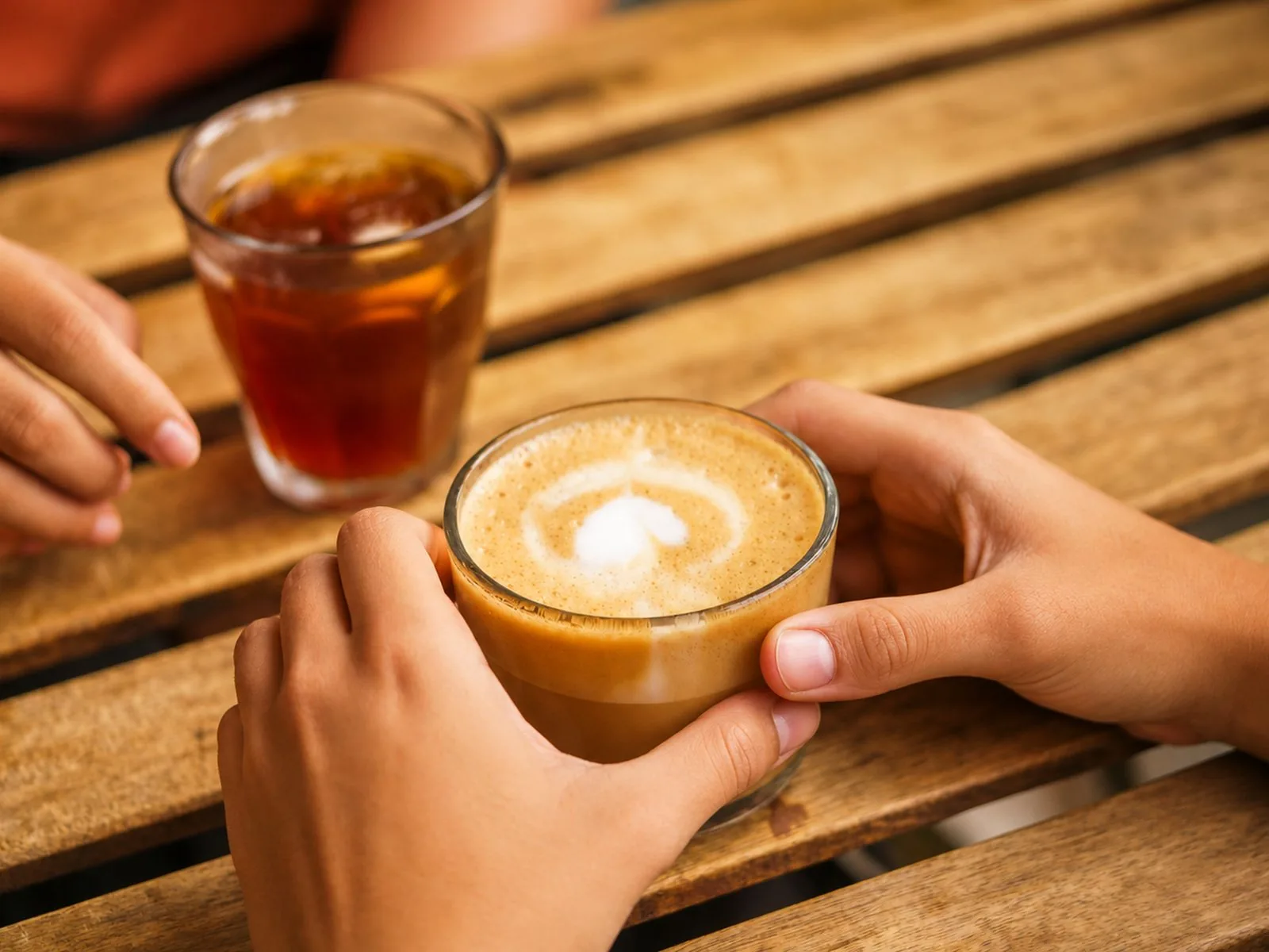 Duas pessoas sentadas a uma mesa de madeira ao ar livre, uma delas a segurar uma taça de café com leite